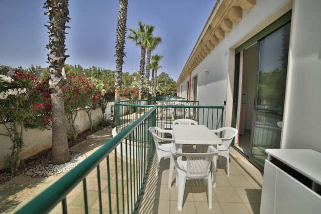 Un balcone all'aperto con tavolo e sedie bianche, circondato da palme e piante fiorite.