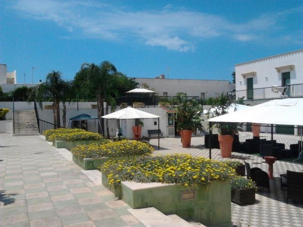 Cortile soleggiato con sedie bianche, ombrelloni e piante, tra edifici bianchi.