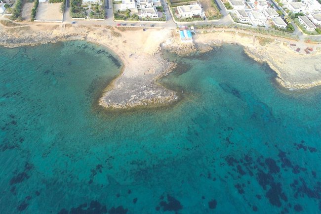 Veduta aerea di una costa rocciosa con acque blu e alcuni edifici sullo sfondo.