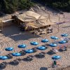 Ombrelloni blu ordinati su una spiaggia dall'alto, davanti a una struttura con tetto di paglia.