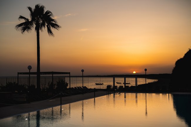 Tramonto sul mare con piscina in primo piano e una palma.