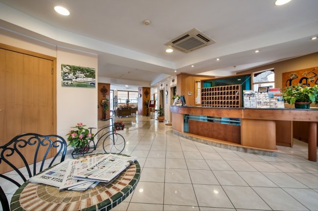 Reception di un hotel con bancone in legno, tavolo con giornali e decorazioni floreali.