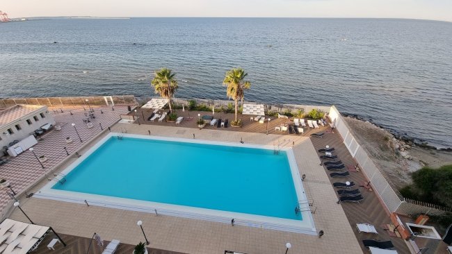 Vista dall'alto di una piscina rettangolare vicino alla costa, con sedie a sdraio e palme.