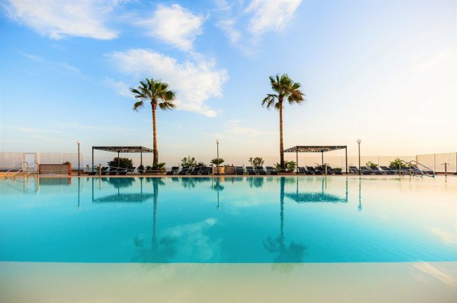 Piscina all'aperto con due palme e cielo azzurro riflesso nell'acqua.