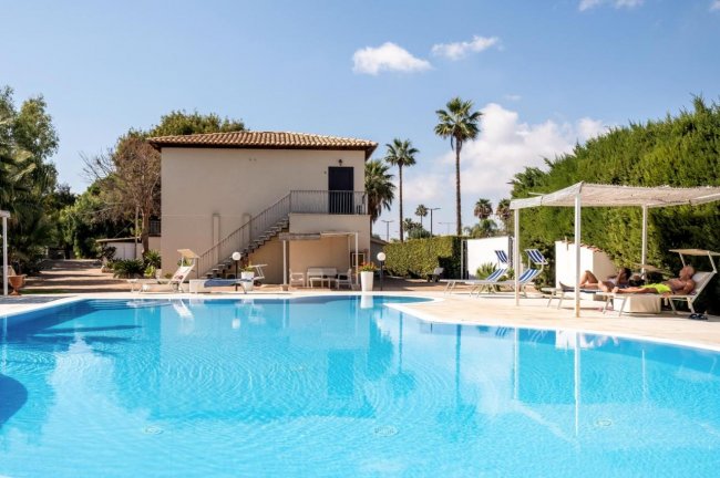 Piscina con sdraio e un edificio a due piani sullo sfondo.