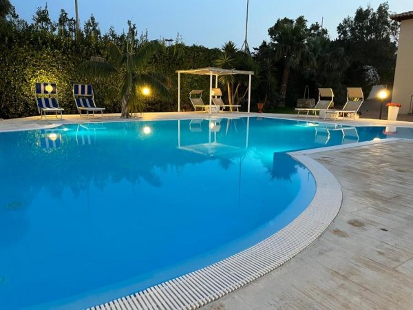 Piscina all'aperto illuminata di sera, circondata da lettini e vegetazione.