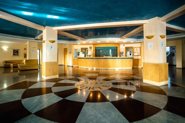 Ampia hall con pavimento decorato a raggi, bancone reception, divani, soffitto blu e colonne beige.
