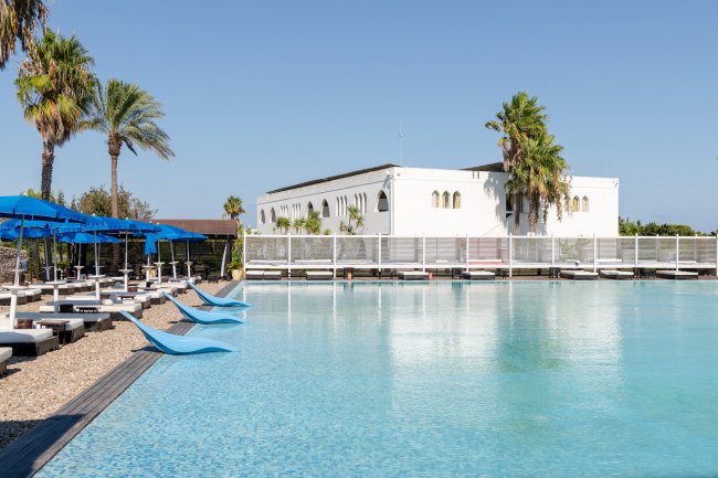 Piscina con sedie a sdraio e ombrelloni blu, edificio bianco e palme sullo sfondo.