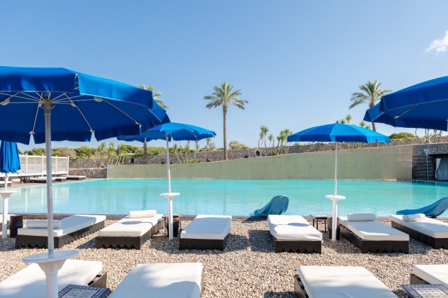 Lettini e ombrelloni blu lungo il bordo di una piscina, palme e cielo sereno.