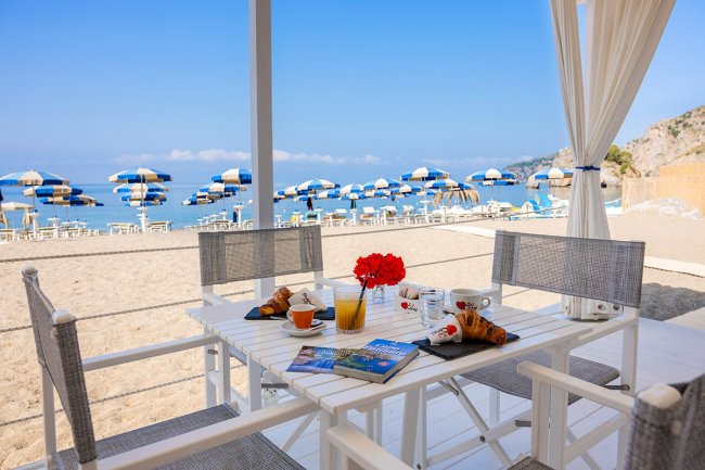 Tavolo all'aperto con colazione vista mare e ombrelloni blu in spiaggia.