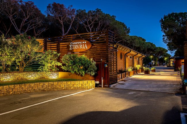Ingresso Camping Etruria illuminato con vegetazione.