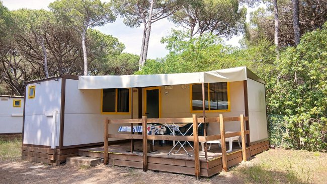 Casa mobile con terrazza in legno, tavolo, sedie, tende. Circondata da alberi alti e vegetazione.