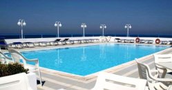 Piscina all'aperto con lettini e lampioni sullo sfondo di un cielo blu chiaro.