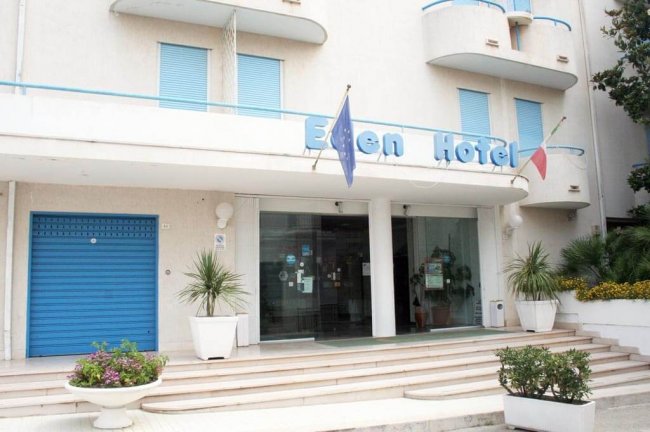 Facciata di un hotel con ingresso a vetri, scalini e bandiere europee.