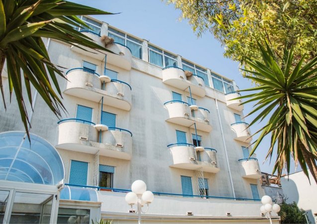 Edificio a più piani con balconi curvi e pareti bianche, circondato da alberi.