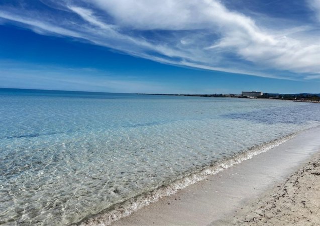 Spiaggia con sabbia chiara, mare cristallino e cielo azzurro parzialmente nuvoloso.