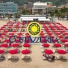 Spiaggia con ombrelloni rossi e un logo giallo e blu al centro.