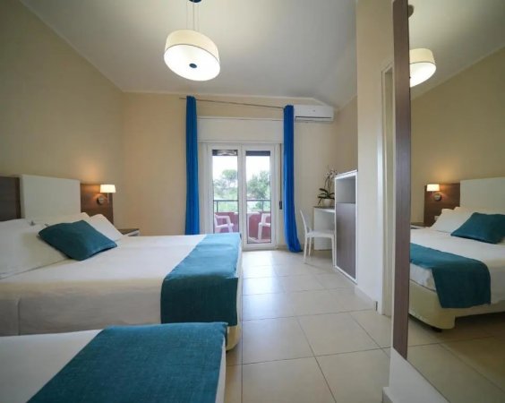 Stanza d'albergo con due letti, coperte e cuscini blu, porta-finestra con tende blu.