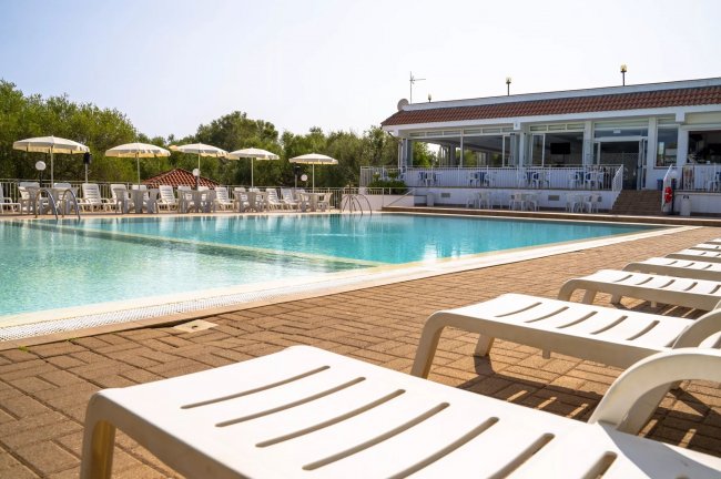 Piscina con bordo in legno, affiancata da lettini e una struttura bianca sullo sfondo.