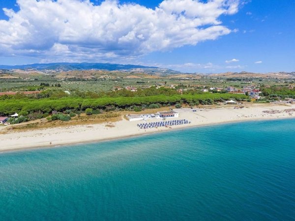 Spiaggia con lettini e ombrelloni e mare azzurro. Vegetazione e colline sullo sfondo.
