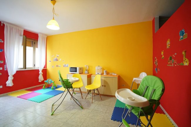 Stanza colorata con pareti gialle e rosse, sedie colorate, tavolino e seggioloni per bambini.