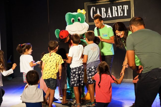 Bambini circondano una mascotte verde su un palco con la scritta 