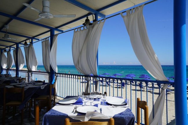 Tavoli apparecchiati con vista sul mare, tende bianche sventolano sotto una tettoia azzurra.