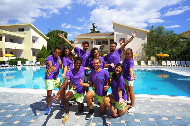 Gruppo di persone davanti a una piscina, indossano magliette viola e pantaloncini verdi.