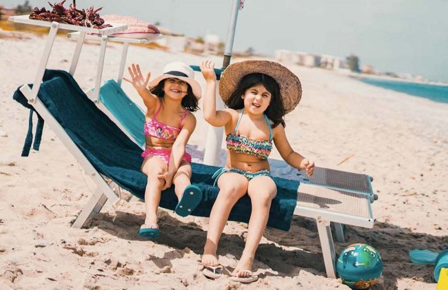 Due bambine in costume da bagno salutano sulla spiaggia, sedute su lettini con cappelli estivi.