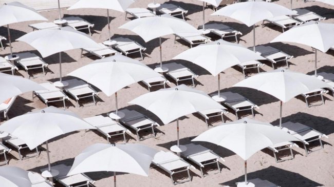 Spiaggia con file di lettini e ombrelloni bianchi disposti geometricamente.